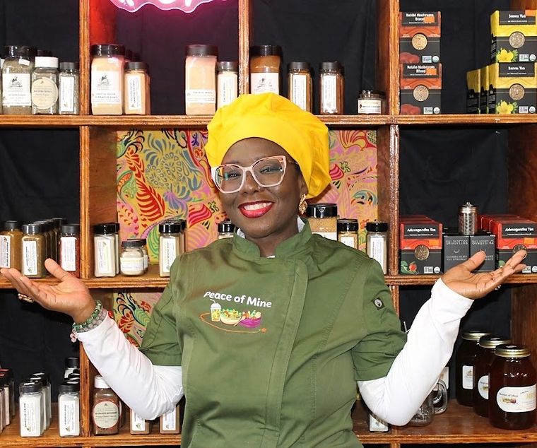 Chef Tatiana Winters in her Peace of Mine chef coat, arms open in front of the shop shelves