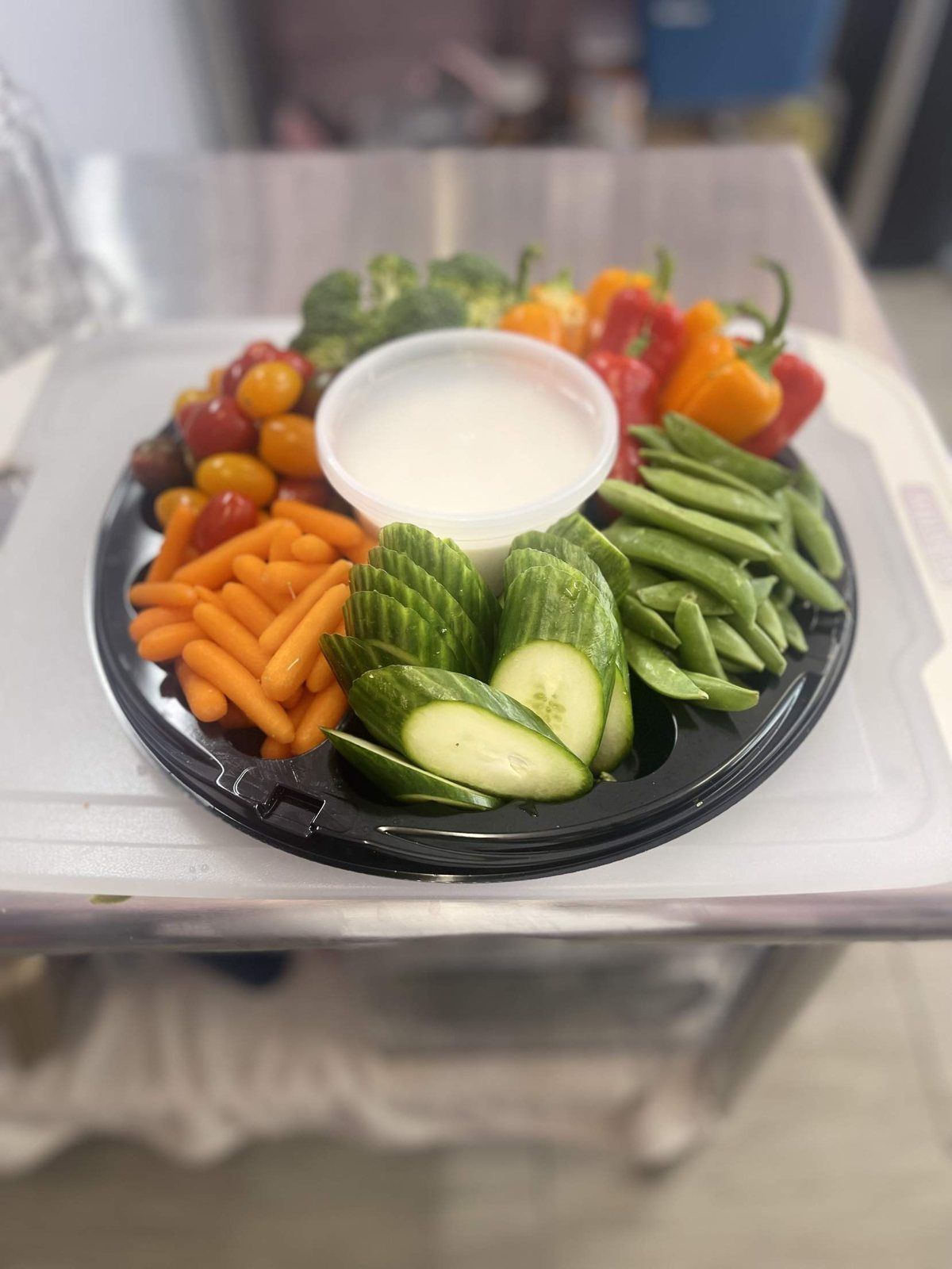 A round tray of carrots, cucumber, snap peas, peppers, broccoli and tomatoes around dip